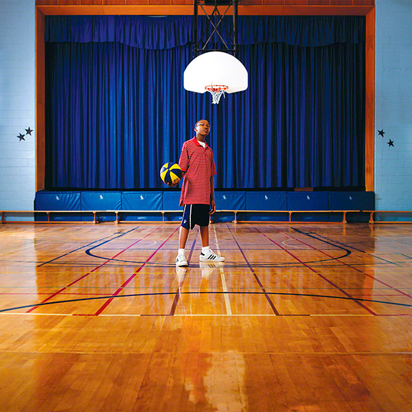 dynamic portrait of a black student posing with a basketball on interior court, middle school gym with stage in background interior