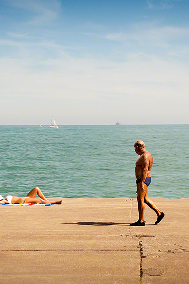 old man in speedo checking out young woman sunbathing near water front
